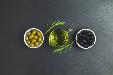 Tabaklar, zeytin yağı ve biberiye siyah bir taş zemin üzerine siyah ve yeşil zeytin ayarlayın. Üstten görünüm, kopya alanı.