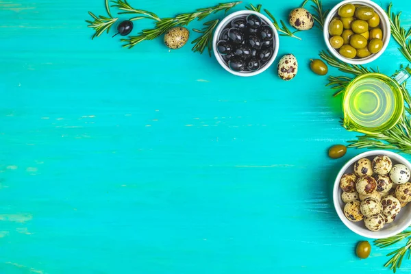 Set of black and green olives, quail eggs on plates, olive oil and rosemary, on a blue turquoise wooden table background. Top view, copy space.