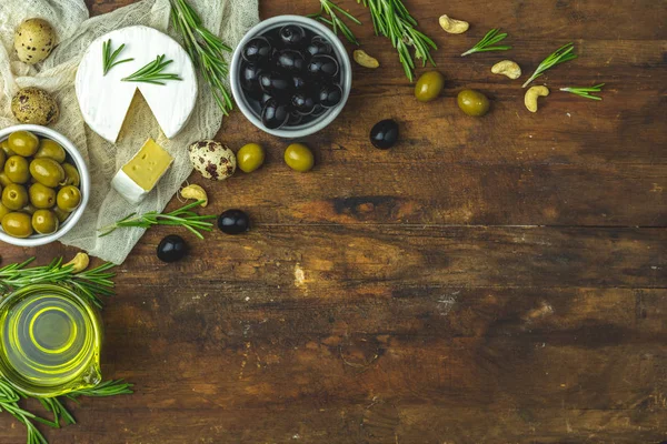Set of cheese camembert, black and green olives, quail eggs on plates, olive oil and rosemary, cashew nuts, on old dark wooden table background. Top view, copy space.