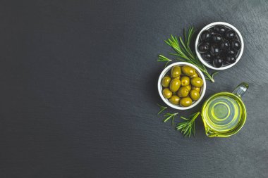 Tabaklar, zeytin yağı ve biberiye siyah bir taş zemin üzerine siyah ve yeşil zeytin ayarlayın. Üstten görünüm, kopya alanı.