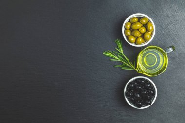 Tabaklar, zeytin yağı ve biberiye siyah bir taş zemin üzerine siyah ve yeşil zeytin ayarlayın. Üstten görünüm, kopya alanı.