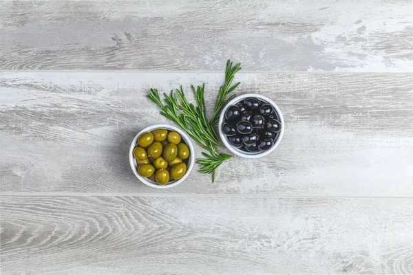 Set of black and green olives on plates and rosemary on a light gray wooden background. Top view, copy space.