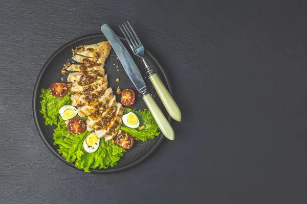Delicious salad, chicken with honey and mustard souse, lettuce, quail eggs, cherry tomatoes and sesame in black ceramic plate, top view, copy space, hawaiian, asian food.