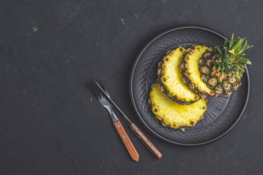 Ananas dilimlenmiş seramik tabak ve çatal bıçak seti, çatal siyah taş beton yüzey arka plan dokulu. Metniniz için kopya alanı ile üstten görünüm.