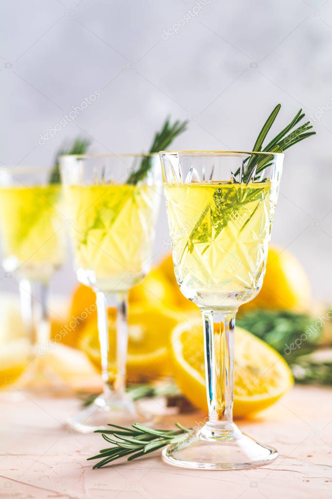 Traditional italian homemade lemon alcohol drink liqueur limoncello with pieces of lemon and rosemary herb on light pink, peach or coral color stone concrete surface