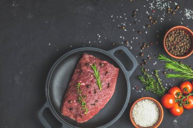 Taze çiğ et Dana biftek siyah tavada pişirme baharat, kekik, biberiye ve domates koyu arka plan, üstten görünüm için malzemelerle kopya alanı sizin için metin.