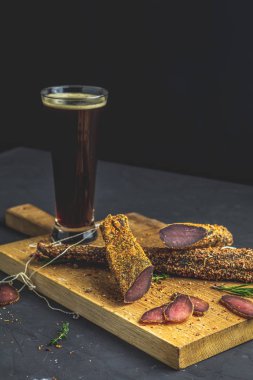 Karanlık bira cam ve sarsıntılı, basturma, kurutulmuş et sığır eti, et eti ile baharat ahşap kesme tahtası, siyah Beton yüzey tablo arka plan üzerinde füme.