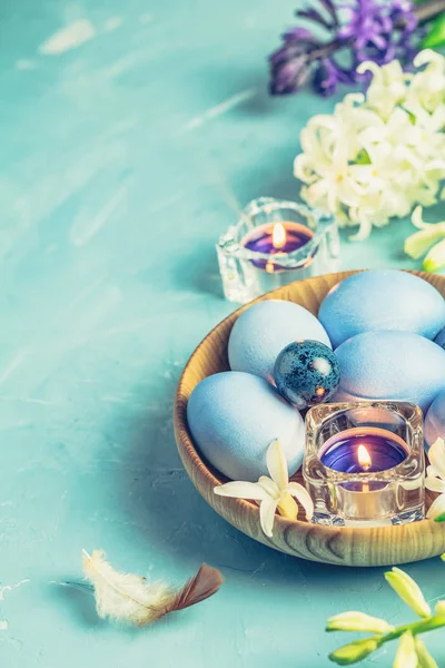 Easter greeting card with colored blue eggs, quail eggs and candles  in wooden plate in front of white and blue hyacinths over blue concrete surface background. Happy Easter festive background