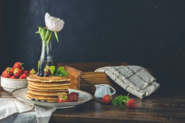 Srtawberry, yaban mersini ve naneli krep seramik tabakta, küçük seramik kavanozdan şurup ve koyu ahşap bir masa üzerinde çiçekler ve siyah arkaplan. 