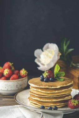 Srtawberry, yaban mersini ve naneli krep seramik tabakta, küçük seramik kavanozdan şurup ve koyu ahşap bir masa üzerinde çiçekler ve siyah arkaplan. 
