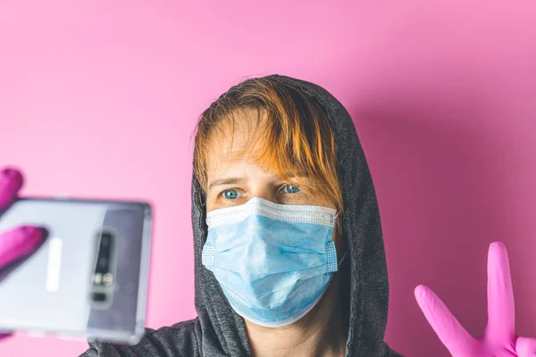 Tired female medical volunteer in a medical mask and hood take a selfie on a pink background. Health protection during flu virus outbreak, coronavirus epidemic infectious diseases