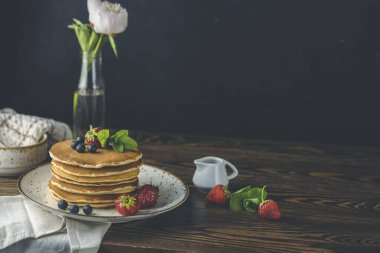 Çilekli krep, seramik tabakta yaban mersini ve nane şekeri, küçük seramik kavanozdan şurup ve koyu ahşap bir masada çiçekler.