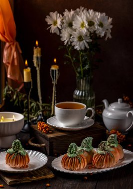 Balkabağı şekilli kekler mum, çiçek ve deniz dikenli böğürtlenlerle süslenmiş bir masada çay ile servis edilir ve sonbahar şenlikleri için samimi bir atmosfer yaratır.