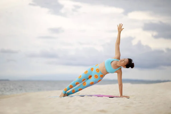 asian woman playing side plank yoga pose on white sand beach