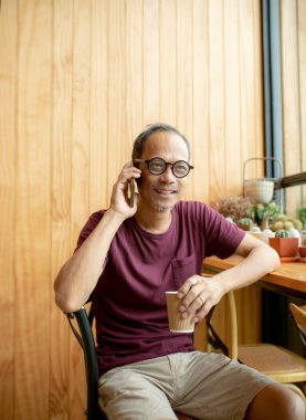 Asyalı adam sol elinde kahve fincanıyla akıllı telefondan konuşuyor.