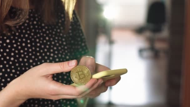 Kvinna med en smartphone med en hand och hålla ett bitcoin mynt med den andra. Bitcoin är en världsomspännande kryptovaluta och digitala betalningssystem. — Stockvideo