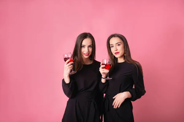 两个漂亮的双胞胎女孩庆祝企业新年派对 喝酒聊天 粉红的背景 明亮的妆容 身穿黑色时髦晚礼服的女孩 — 图库照片