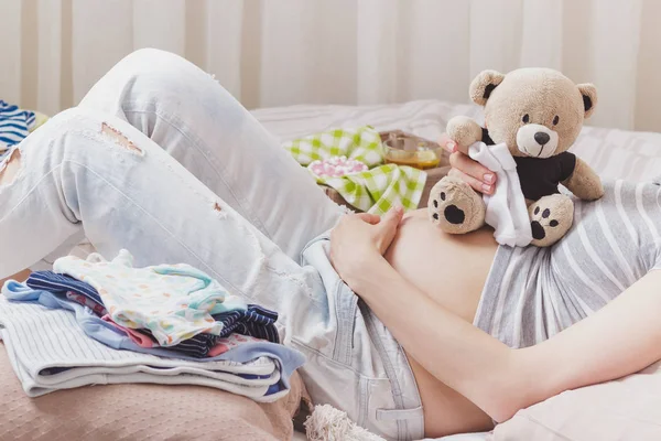 Pregnant woman with a teddy bear and various clothes for a newborn, top view