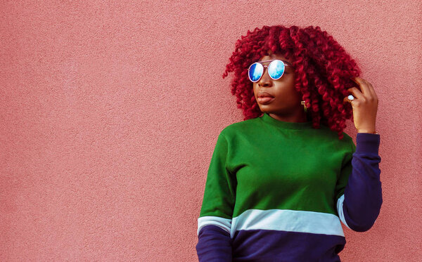 Charming young African female in blue sunglasses. Fashionable and stylish girl posing near pink wall