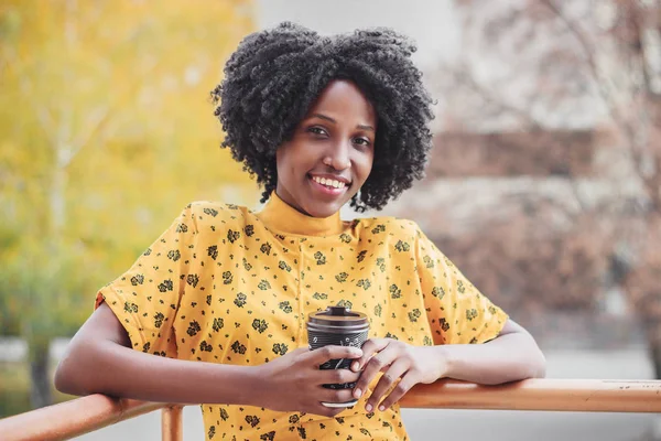 Young dark-skinned woman dressed in casual clothes holding cup of hot drink, enjoying coffee