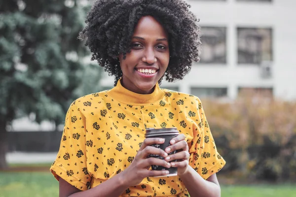 Young dark-skinned woman dressed in casual clothes holding cup of hot drink, enjoying coffee