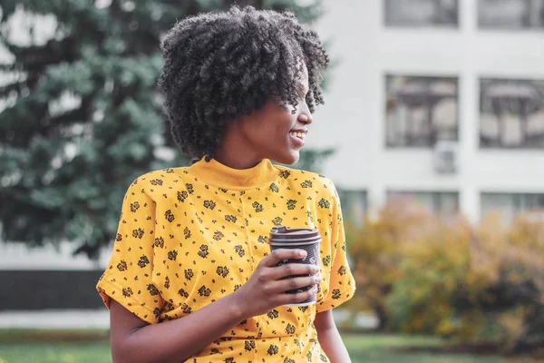 Young dark-skinned woman dressed in casual clothes holding cup of hot drink, enjoying coffee