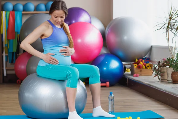 Pregnant woman sits on the fitness ball in the gym, soft focus background