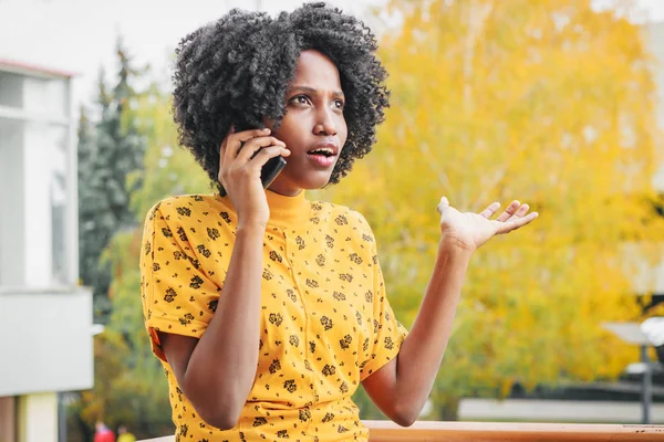 Young smiling dark-skinned woman dressed in casual clothes talking by her phone, blurred background