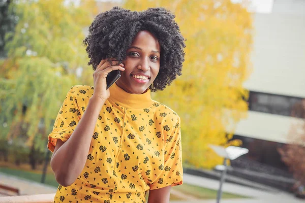 Young smiling dark-skinned woman dressed in casual clothes talking by her phone, blurred background
