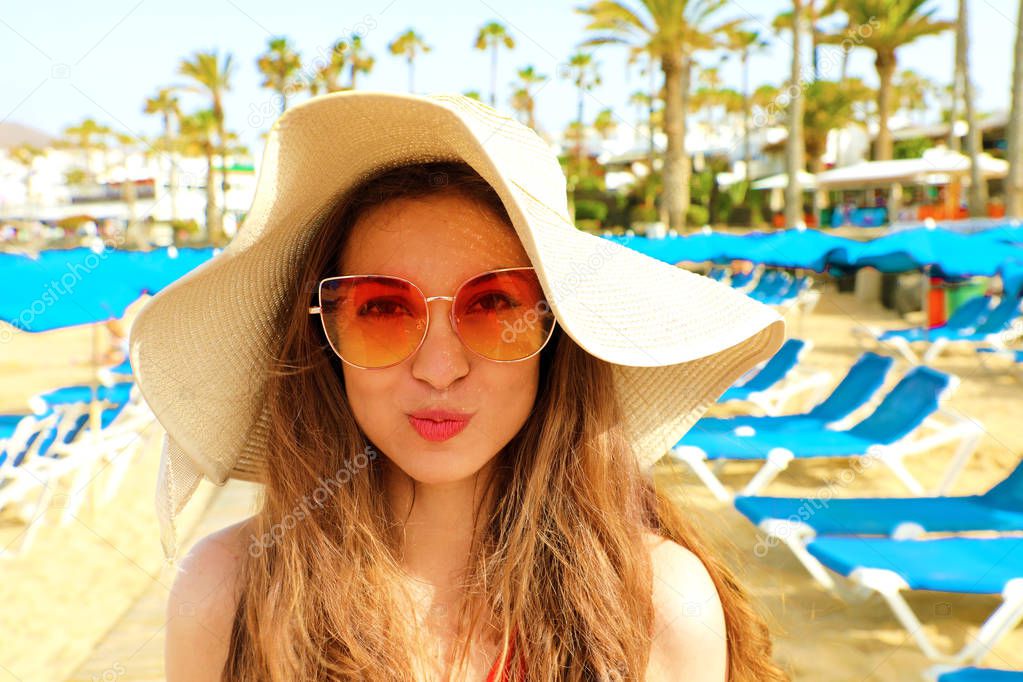Cute blonde woman on the beach, having fun and sending air kiss. Travel in tropical luxury beach vacation, sunglasses, straw hat, pretty face.
