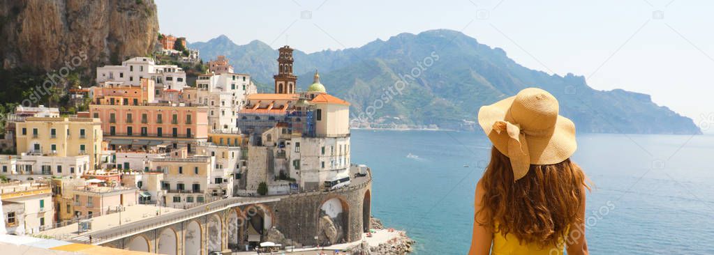 Summer holiday in Italy panorama banner. Back view of young woman with straw hat and yellow dress with Atrani village on the background, Amalfi Coast, Italy.