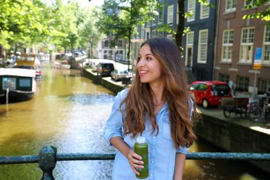 Kentsel şehir yaşam tarzı hippi kız içecekler yeşil suyu Amsterdam kanal, Hollanda. Sağlıklı detoks vegan diyet sebze soğuk preslenmiş ile pürüzsüz için ayıklayın. Vegan yemek ve detoks mola zaman kavramı.