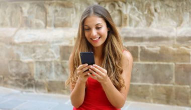 Gülümseyen kadın video oyunları oynamak için cep telefonu app kullanarak. Şehir kadın rahatlatıcı. Kentsel yaşam tarzı. Afiş kırpma için reklam kopya alanı, arka plan Avrupa eski kasaba taş duvar. 