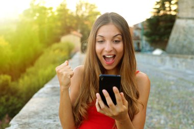 Mutlu genç kadın parlama ile sokakta yürürken akıllı telefonunu izliyor. Açık havada akıllı telefon kullanan sürpriz kız.