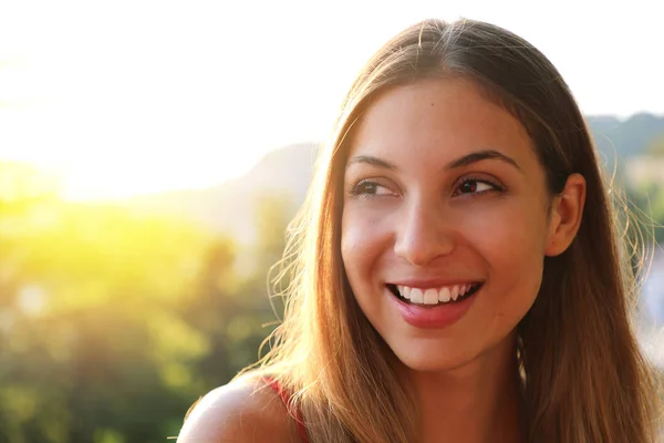 Woman smiling with perfect smile and white teeth thinking and looking sideways in park in summer. Sunlight flare. Copy space.