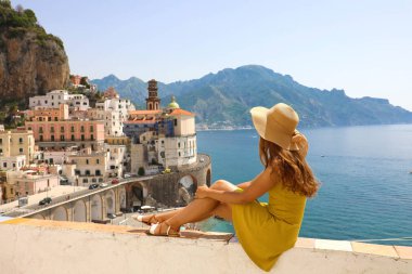 Duvarda oturan şapkalı güzel genç kadın İtalya 'nın Amalfi Sahili' ndeki çarpıcı panoramik köy Atrani 'ye bakıyor.