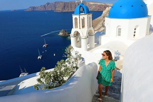 Santorini fashion woman climbing steps in Oia, famous white village with blue domes in Greece. Girl in green dress and sunglasses looking to the side faboulos landscape with blue Mediterranean sea.