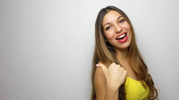 Laughing young woman in yellow tank top looking at the camera and pointing away with thumb over white background