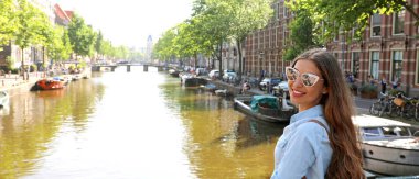 Güneş gözlüğü ve Amsterdam şehir zevk sırt çantası ile seyahat kız portresi. Genç kadın kameraya Amsterdam kanal, Hollanda, Avrupa arıyor görünümünü panoramik afiş.
