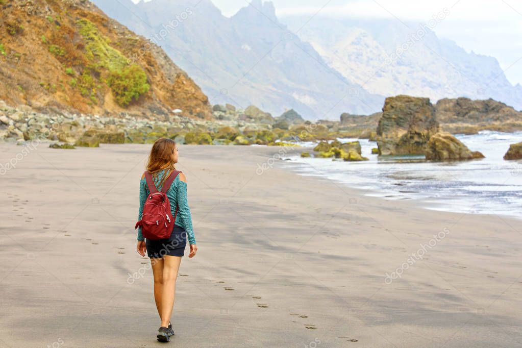 Young female hiker discovering wild paradisiac beach in Tenerife Island. Back view traveler girl arrives on hidden amazing beach of Canary Islands, Spain.