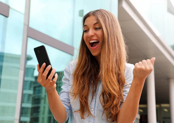 Eufórica mujer de negocios viendo su teléfono inteligente y mostrando el gesto ganador al aire libre —  Fotos de Stock