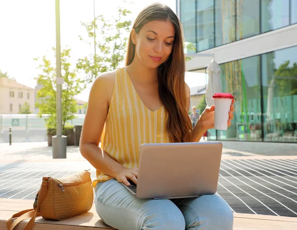 Όμορφη νεαρή γυναίκα που εργάζεται στο laptop καθιστό υπαίθριο κρατώντας φλιτζάνι καφέ. — Φωτογραφία Αρχείου