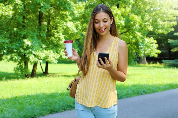 Mulher bonita mensagens no telefone inteligente e andando no parque com café na mão . — Fotografia de Stock