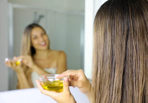 Mujer joven que aplica mascarilla de aceite de oliva en el cabello delante de un espejo. Concepto de cuidado del cabello. Céntrate en el cabello . — Foto de Stock