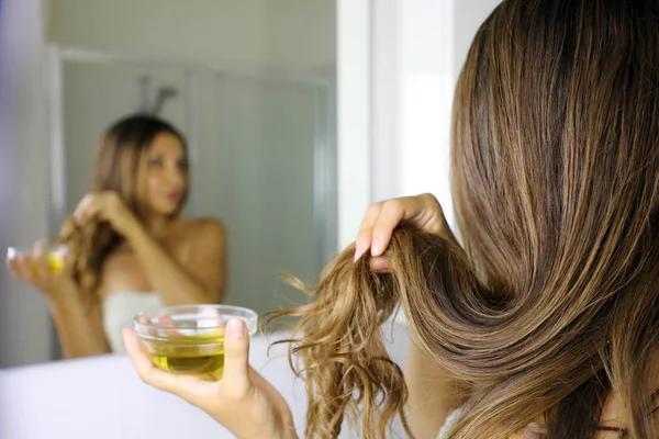 Junge Frau, die vor einem Spiegel eine Olivenölmaske auf Haarspitzen aufträgt. Haarpflegekonzept. Fokus auf Haare. — Stockfoto