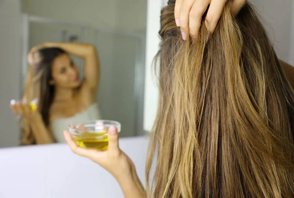 Junge Frau, die vor einem Spiegel Olivenölmaske auf die Haare aufträgt. Haarpflegekonzept. Fokus auf Haare. — Stockfoto