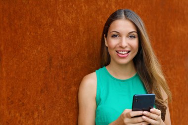 Pas arka planda kameraya bakan akıllı telefon kullanan genç kadın.