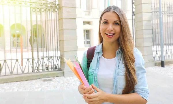 Smiling student girl holding folders with school on the background. Looks at camera. Copy space. — Stock Photo, Image