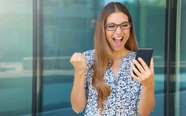 Emocionada joven mujer de negocios que recibe buenas noticias en el teléfono inteligente celebrando con el puño en alto al aire libre. Copiar espacio . — Foto de Stock