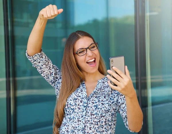 Éxito joven mujer de negocios levanta el brazo con el teléfono inteligente al aire libre . —  Fotos de Stock